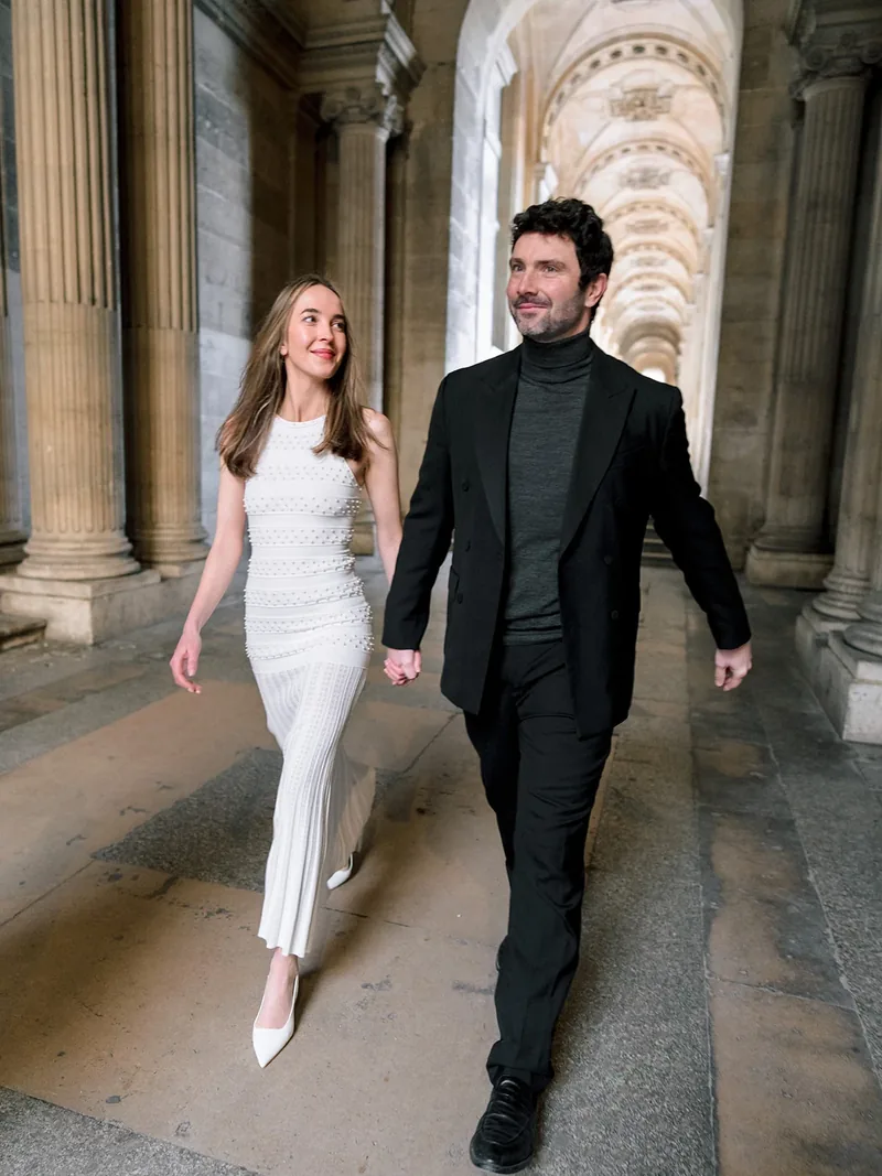 Couple in black and white outfits walking beneath the Louvre colonnade