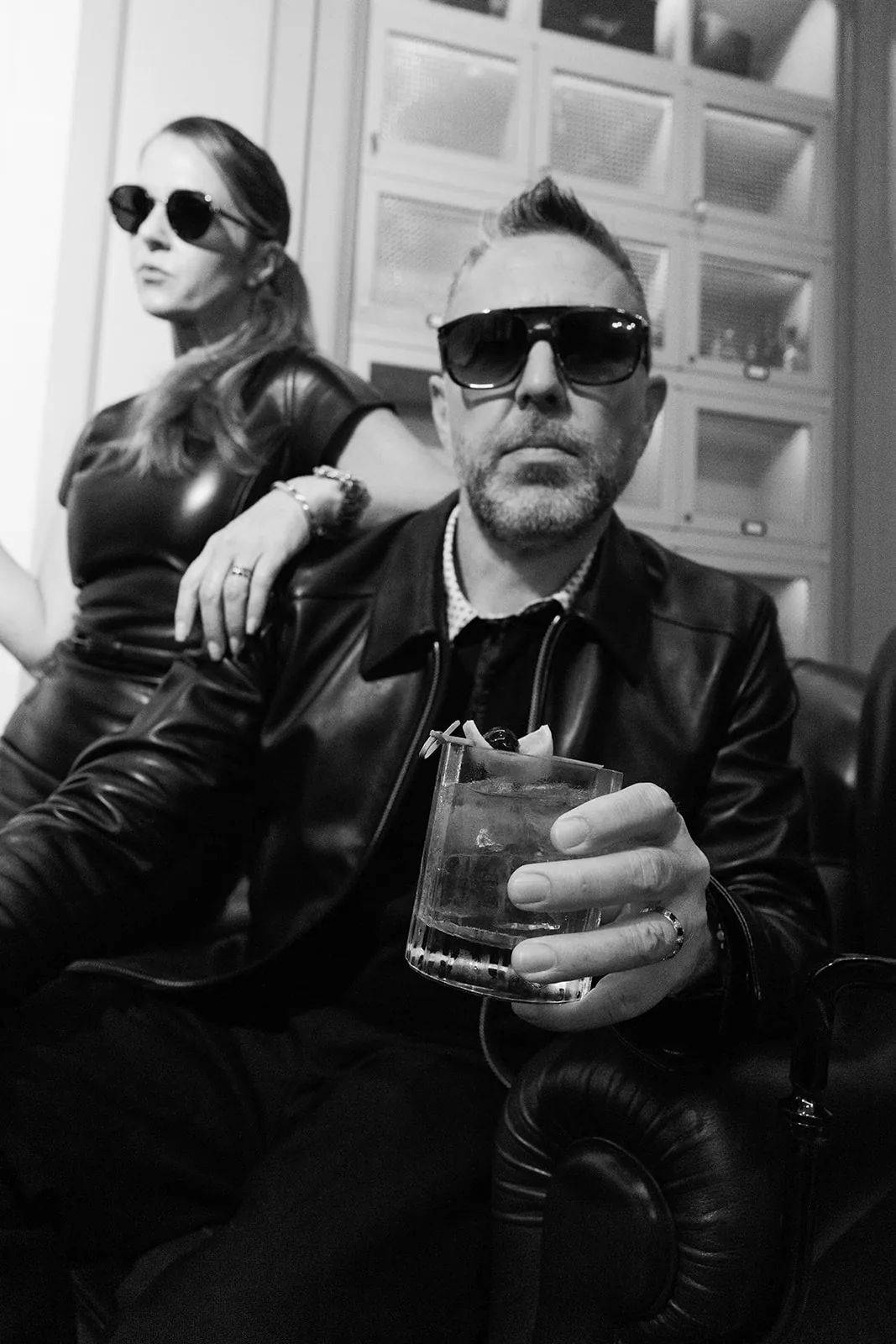 Portrait en noir et blanc d’un homme tenant un cocktail, avec une femme en tenue de cuir derrière lui