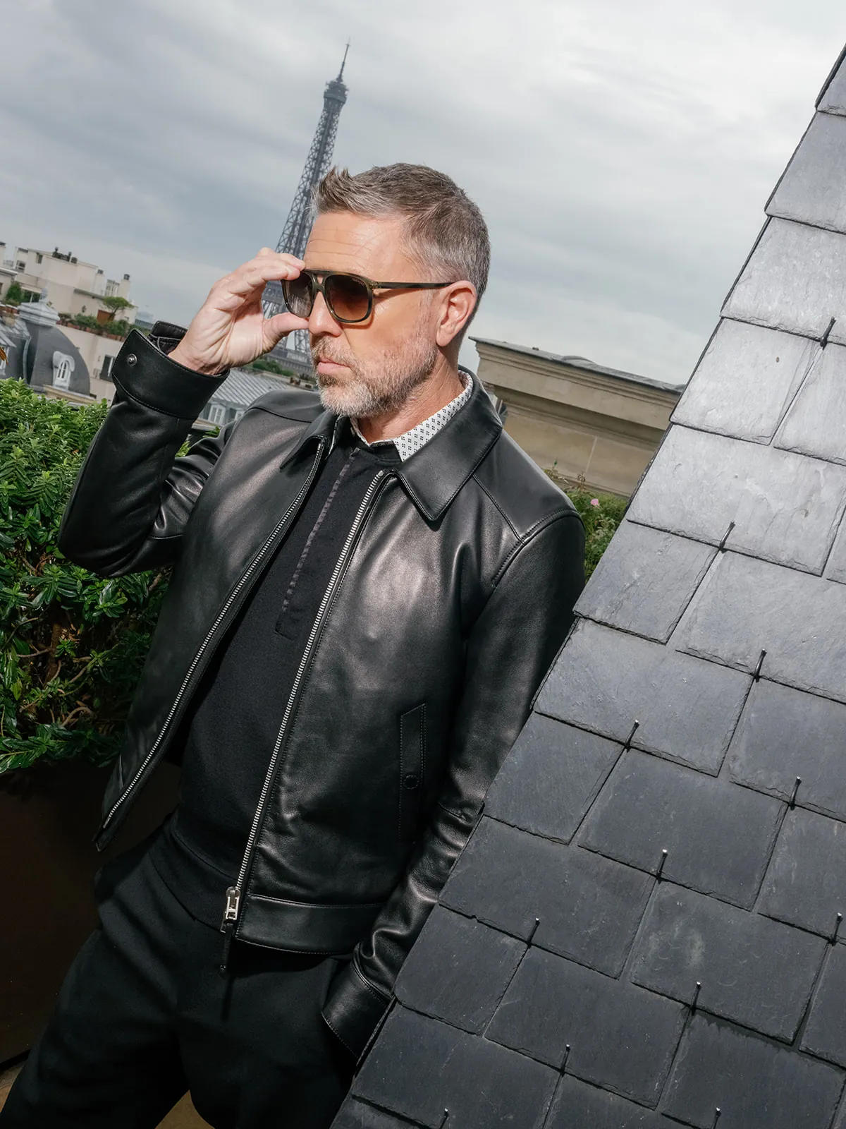 Man adjusting his sunglasses on a Paris rooftop, wearing a black leather jacket