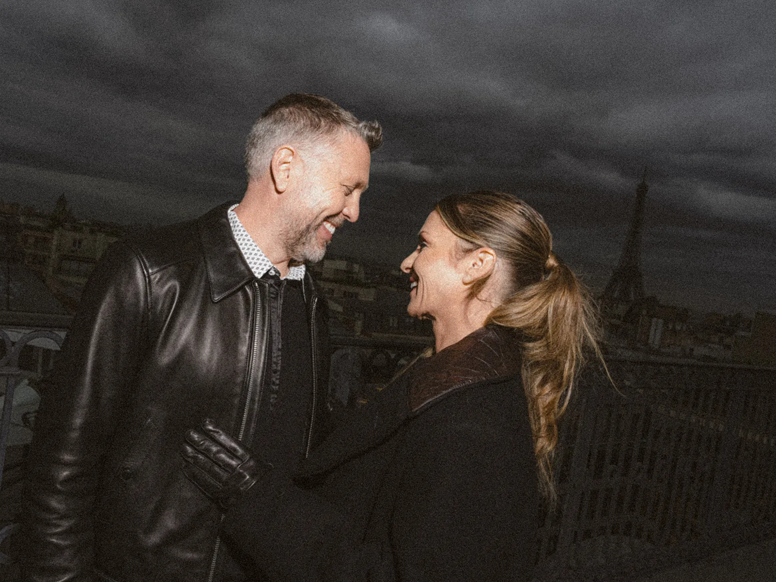 Two people smiling at one another on a Paris rooftop at dusk