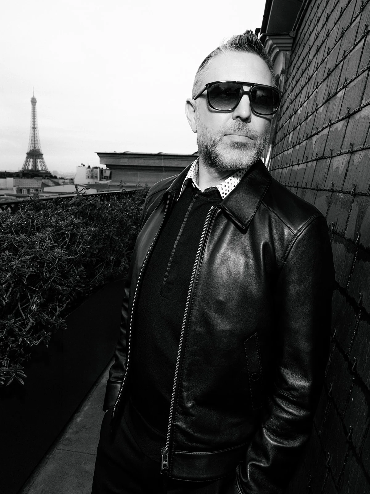 Black and white portrait of a man wearing sunglasses and a black leather jacket, with the Eiffel Tower behind him