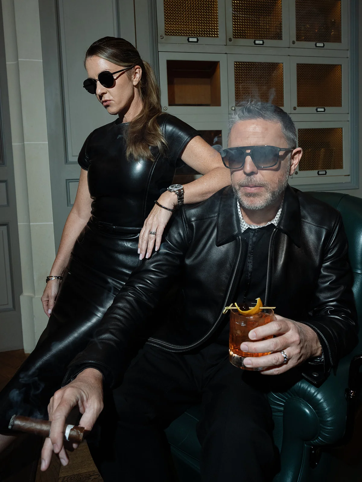Couple wearing black leather outfits and sunglasses beside a green armchair, with a cocktail and cigar